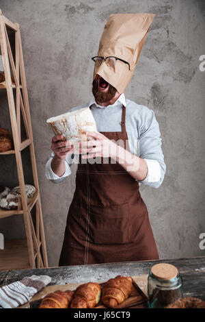 Image of young screaming man baker standing with paper bag on head wearing glasses at bakery holding bread. Stock Photo