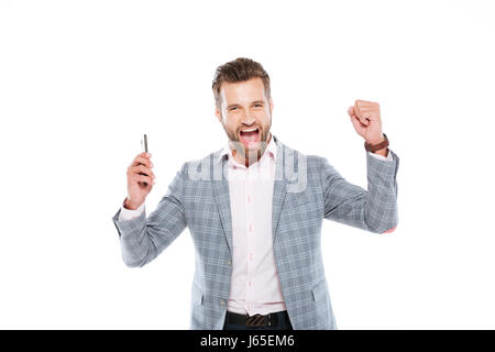 Picture of smiling young man standing isolated over white background and using mobile phone. Looking at camera make winner gesture. Stock Photo