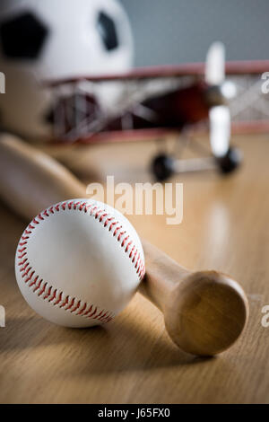Baseball bat and hardball on wooden floor with toy plane and soccer ball on background. Stock Photo