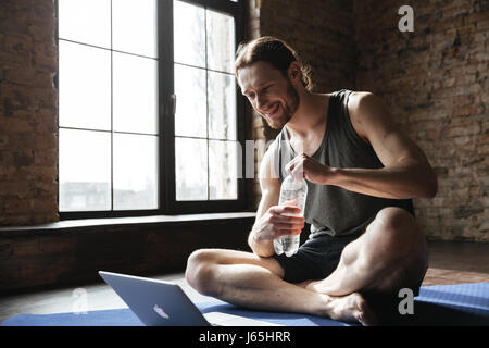 Picture of happy strong sportsman drinking water on floor in gym using laptop computer. Looking aside. Stock Photo
