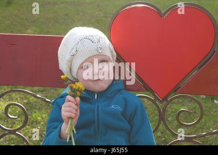 beautiful blonde girl with smiling face with bouquet of yellow dandelion flowers rest under red heart send all love and peace Stock Photo