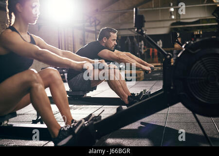 Side view of man and woman doing exercises with rowing machine at gym. Stock Photo