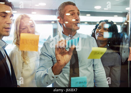 Half body portrait of business manager choosing sticky note on glass in office with colleagues in background, idea concept Stock Photo