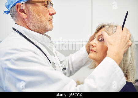 Cosmetology surgeon standing with pencil and holding head preparing senior woman for anti-aging surgery. Stock Photo