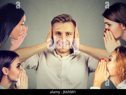 Four women whispering a secret latest gossip to a happy young man who covers ears and ignoring all surrounding noise Stock Photo