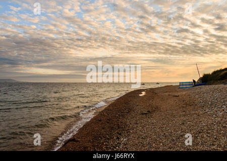 Cloudy sky at sunset on Ringstead Bay, Dorset, UK, a single figure by a backlit multicoloured striped windbreak on the stony beach Stock Photo