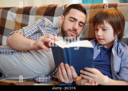 Father and son reading book while relaxing on couch Stock Photo