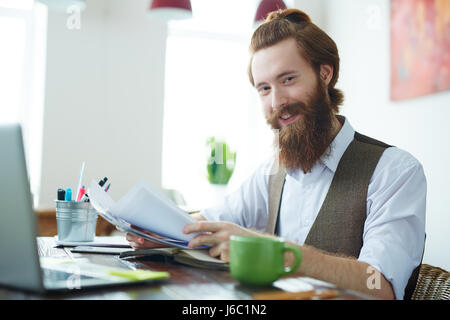 Portrait of trendy bearded man smiling cheerfully, looking at camera, enjoying freelance work from  home office Stock Photo