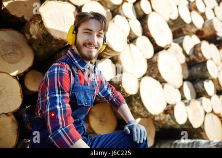 Portrait of man wearing earmuffs looking at camera and smiling while working on timber cutting site, sitting against pile of logs