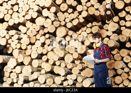 Portrait of young man working on timber cutting site, reading documentation standing against pile of wood logs