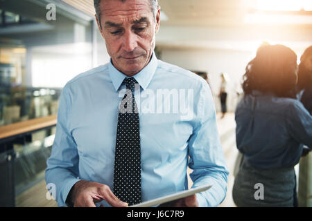 Contemporary mature pensive businessman standing and browsing tablet in office. Stock Photo