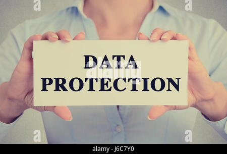 Businesswoman hands holding white card sign with data protection text message isolated on grey wall office background. Retro instagram style image Stock Photo