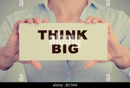 Businesswoman hands holding white card sign with think big text message isolated on grey wall office background. Retro instagram style image Stock Photo