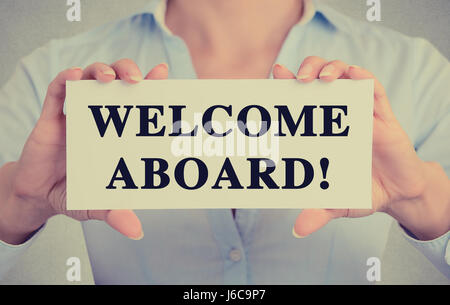 Businesswoman hands holding white card sign with welcome aboard text message isolated on grey wall office background. Retro instagram style image Stock Photo