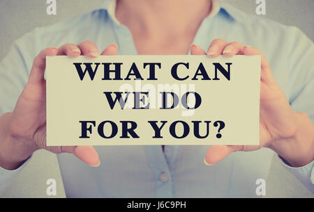 Businesswoman hands holding white card sign with What can we do for you? text message isolated on grey wall office background. Retro instagram style i Stock Photo