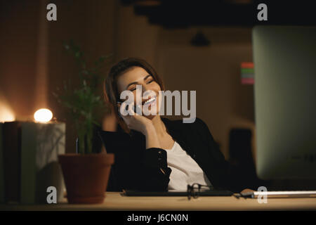 Picture of cheerful young woman designer sitting indoors at night using computer and talking by phone. Looking aside. Stock Photo