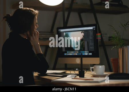 Back view photo of young woman designer sitting indoors at night using computer and talking by phone. Stock Photo