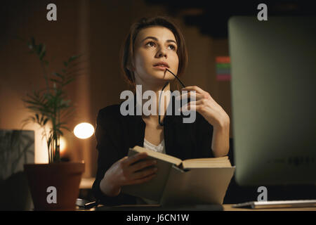 Photo of concentrated young lady designer sitting indoors at night using computer and reading book. Looking aside. Stock Photo