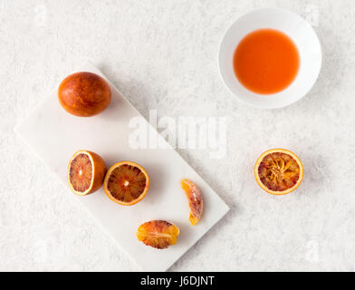 Whole, halved and squeezed blood oranges and fresh juice on white marble chopping board and white textured marbled background with copy space. Stock Photo