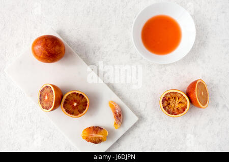 Whole, halved and squeezed blood oranges and fresh juice on white marble chopping board and white textured marbled background with copy space. Stock Photo