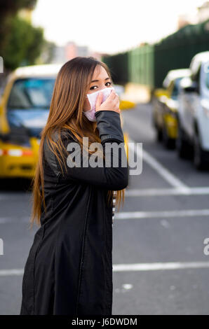 Young woman wearing protective mask and covering her mouth with her hand on the street in the city with air pollution, traffic background Stock Photo