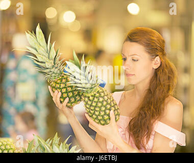 Attractive woman shopping in supermarket. Closeup portrait beautiful young woman picking up, choosing fruits, pineapples in grocery store. Positive fa Stock Photo