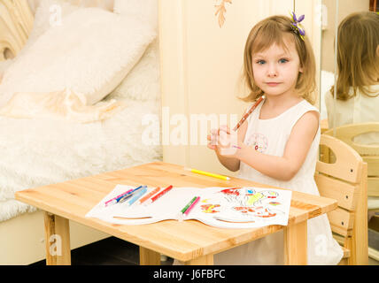 Active little preschool age child, cute toddler girl with blonde curly hair, drawing picture on paper using colorful pencils and felt-tip pens, sitting at wooden table indoors at home or kindergarten Stock Photo