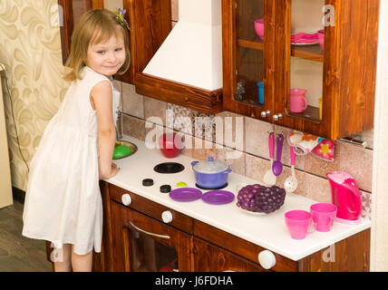 Active little preschool age child, cute toddler girl with blonde curly hair, shows playing kitchen, made of wood, plays in the kitchen Stock Photo