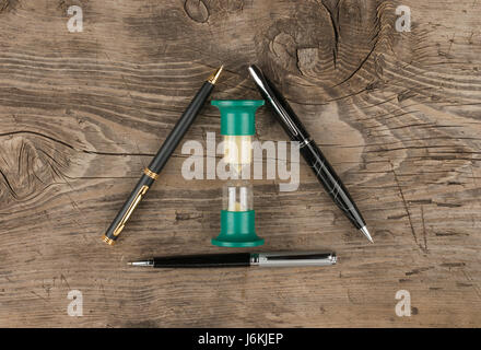 Concept, time and business. Hourglass and three pens on a wooden table. View from above Stock Photo