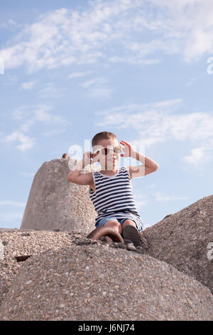 Cute smiling little boy in sunglasses and a vest sitting on a concrete breakwater on blue sky background Stock Photo