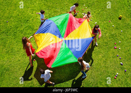 Top view picture of seven kids going round in a circle on the green lawn and holding rainbow parachute Stock Photo