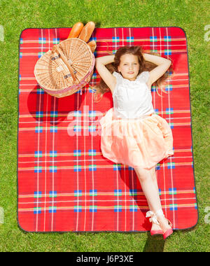Top view portrait of beautiful blond girl in summer clothes relaxing on the red checkered blanket next to the picnic basket Stock Photo