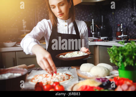 Pretty young woman in apron making pizza in modern kitchen. Stock Photo