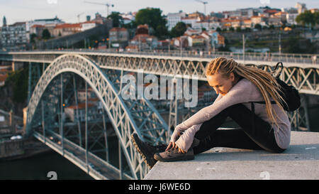 Young woman with blond dreadlocks sitting on the background of Dom Luis I bridge, Porto, Portugal. Stock Photo