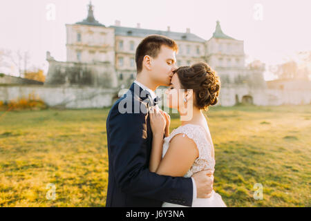 The groom is kissing the beautiful bride in the forehead at the background of the antique gothic castle. during the sunset. Close-up view Stock Photo