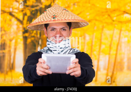 Portrait of a handsome hispanic young business guy wearing an asian conical hat pointing in front of him his Ipad using both hands in autum background Stock Photo
