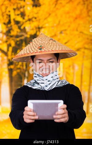 Portrait of a handsome hispanic young business guy wearing an asian conical hat and holding his Ipad in his hands in autum background Stock Photo