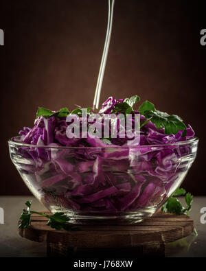 olive oil in a thin stream pour into Salad of red cabbage in glass transparent bowl on dark background Stock Photo