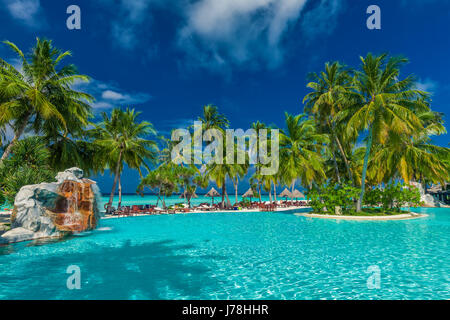 Large infinity swimming pool on the tropical beach with palm trees and umbrellas Stock Photo