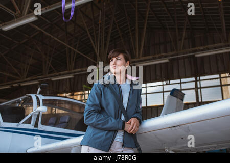 Confident young female pilot leaning on an airplane wing and looking away, she is posing in the airport hangar Stock Photo