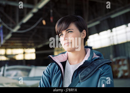 Confident young female pilot posing in the hangar, airplane on the background Stock Photo