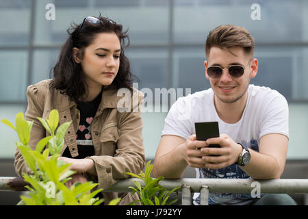 Two young people, students, standing in front of college and looking at mobile phone Stock Photo