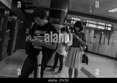 Black & white photo of people standing inside the Tokyo Metro station, with their heads hanging down as they check their mobile phones. Stock Photo