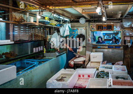 A fishmonger cleaning a fish at the famous Tsukiji Fish Market, one of the largest in the world. Two women sit in an open cubicle in the same stall. Stock Photo