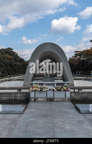 The Hiroshima Cenotaph, framing the A-Dome and the Flame of Peace.  A few people sitting and standing around the Pond of Peace. Stock Photo