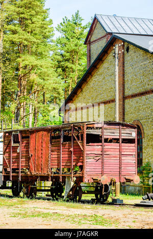 Abandoned red vintage wooden boxcar standing on railway track outside yellow brick building with forest in background. Stock Photo