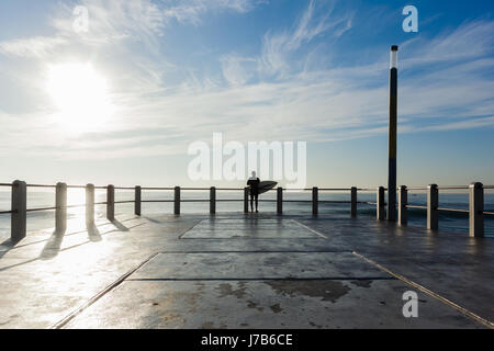 Surfing Surfer silhouetted standing  end of beach ocean pier jetty about to jump into ocean water. Stock Photo