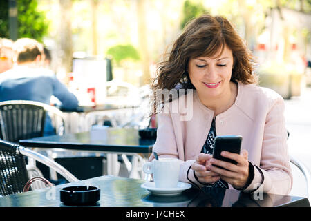 Beautiful and happy middle aged woman having coffee while using a mobile phone Stock Photo