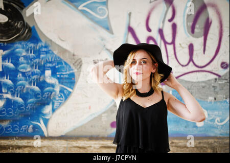 Blonde woman on black dress, necklaces and hat against graffiti wall. Stock Photo