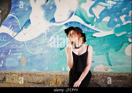 Blonde woman on black dress, necklaces and hat against graffiti wall. Stock Photo